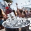 „Flasche FICKEN Likör im Eiskübel mit Eiswürfeln, draußen bei Sonnenschein, im Hintergrund feiernde Menschen