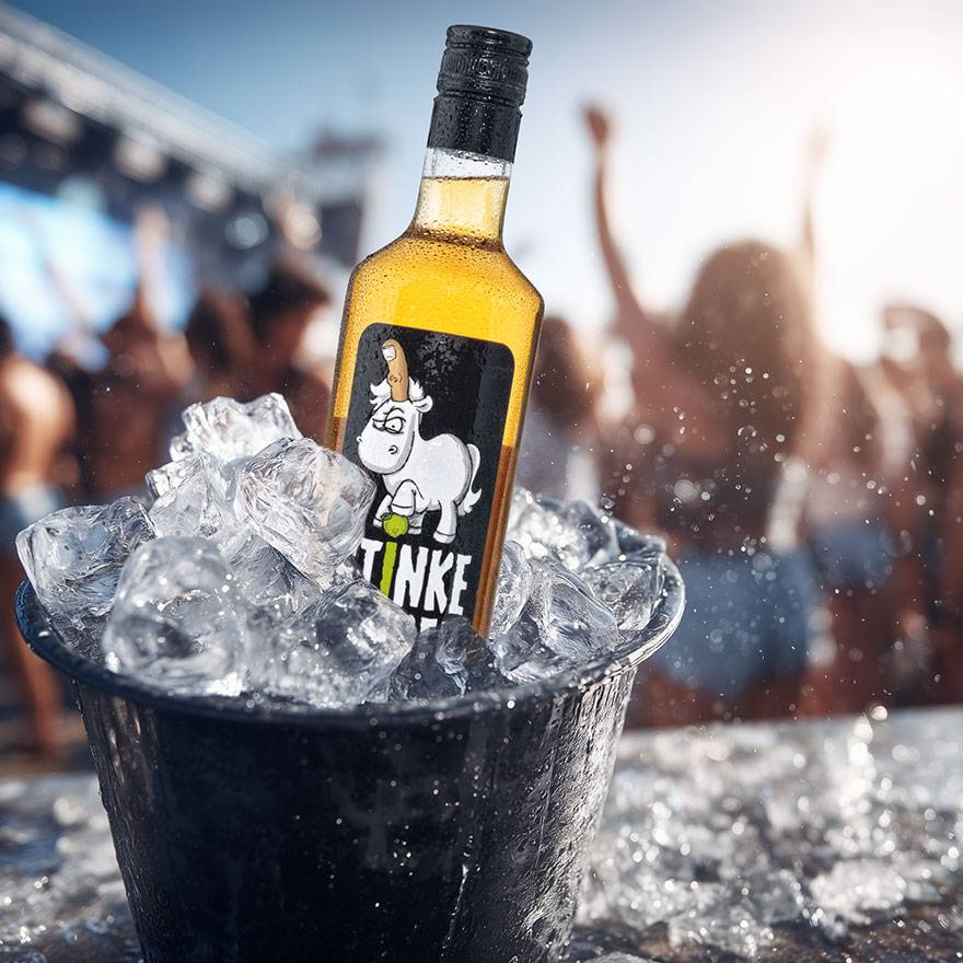 Flasche Stinkefingereinhorn Apfelkorn im schwarzen Eiskübel, umgeben von Eiswürfeln, Partyatmosphäre im Hintergrund.